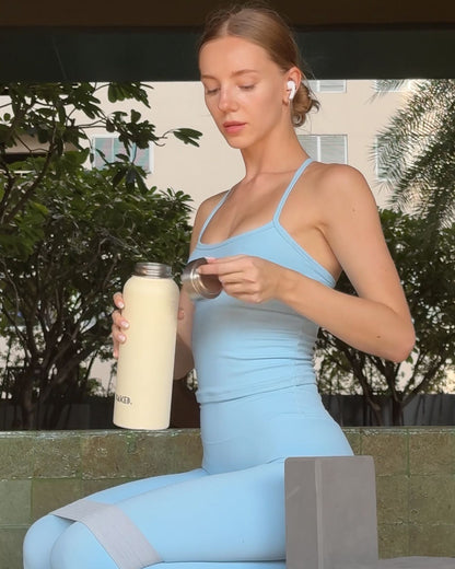 Woman in light blue athletic wear sitting on a mat outdoors, holding a shaker bottle.